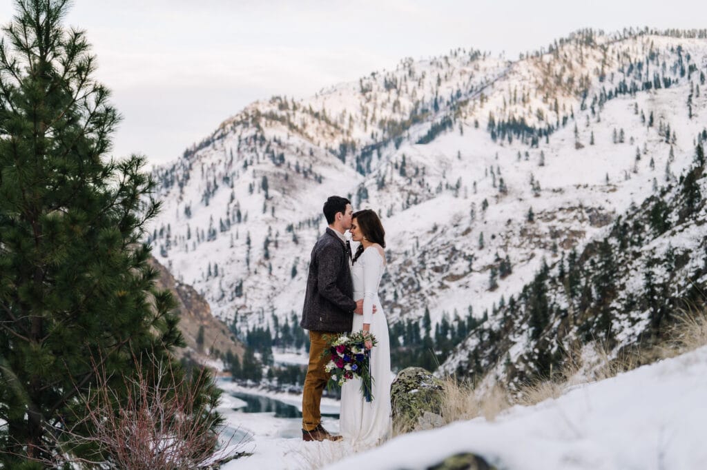 Groom kisses his brides forehead. The couple is standing on a  hill side with the Salmon River canyon behind them. There is snow on the mountains.