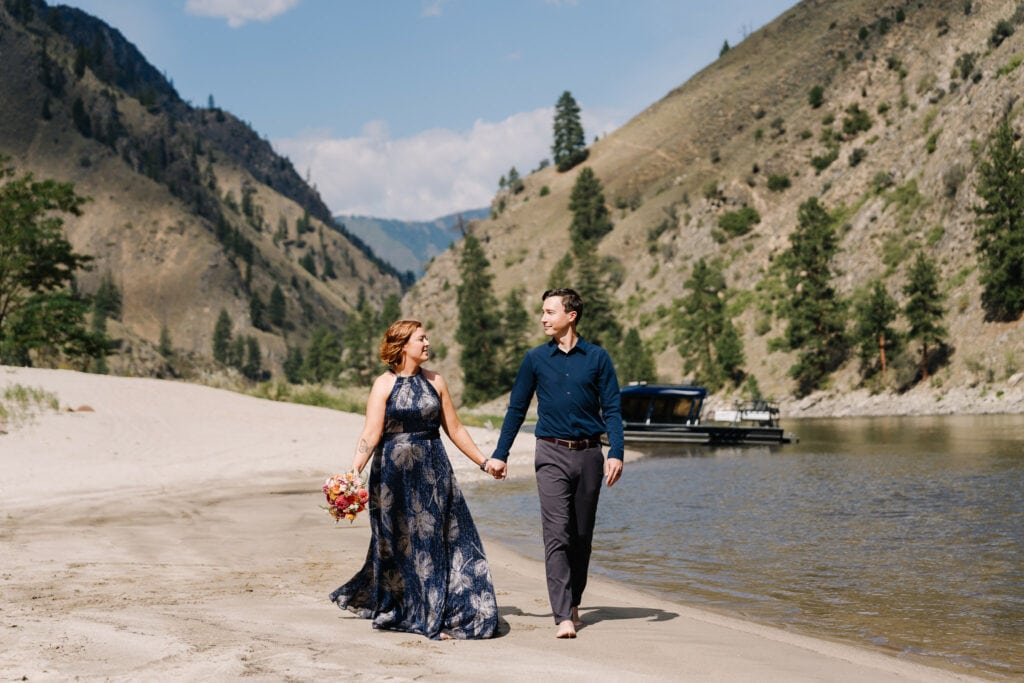 A bride and groom walk down a white sand beach on the Salmon River in Idaho. There is a jet boat in the background. The bride is wearing a blue dress and the groom is wearing a blue shirt and grey dress pants. This couple eloped on the Salmon River in September. 